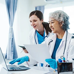 Two female doctors in white lab coats collaborating on a laptop and medical reports, illustrating expertise in the Healthcare & Life Sciences sector.
