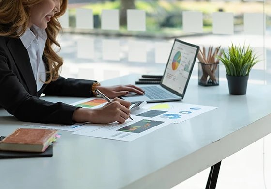 A businesswoman sitting at a desk with a laptop, using a calculator and pen to review financial charts and graphs as part of our ethnography study.