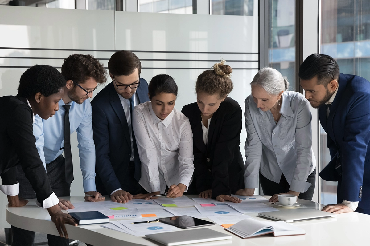 A multiethnic group of male and female business professionals gathered around an office conference table, reviewing charts, graphs, and documents as part of their comprehensive market research methodology.